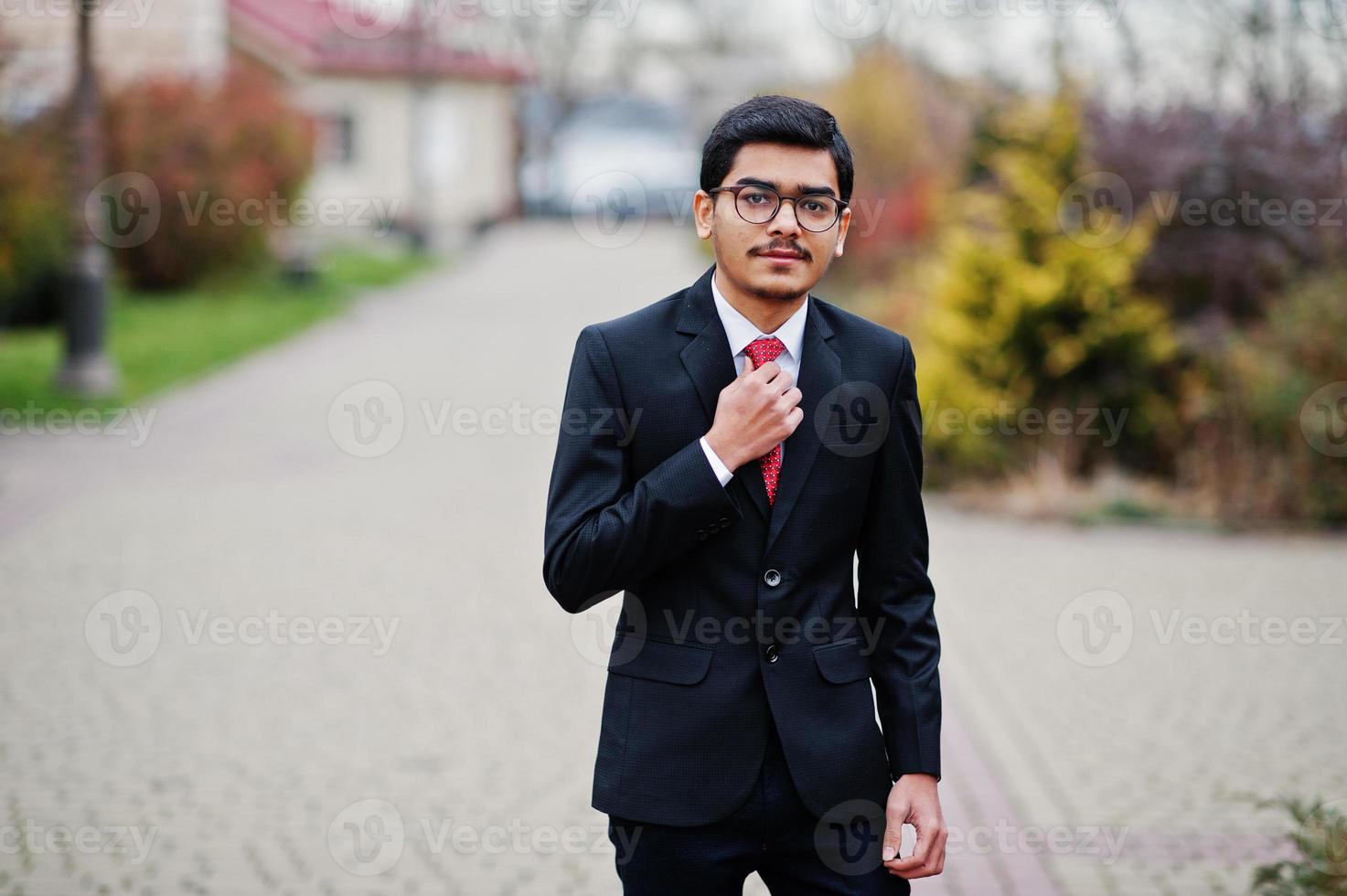 indisk ung man vid glasögon, bär på svart kostym med röd slips poserade utomhus. foto