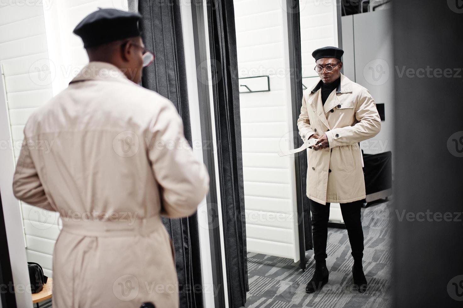 snygg casual afroamerikansk man på svart basker och vit trench-regnrock i provrummet klädaffär, tittar på spegeln. foto