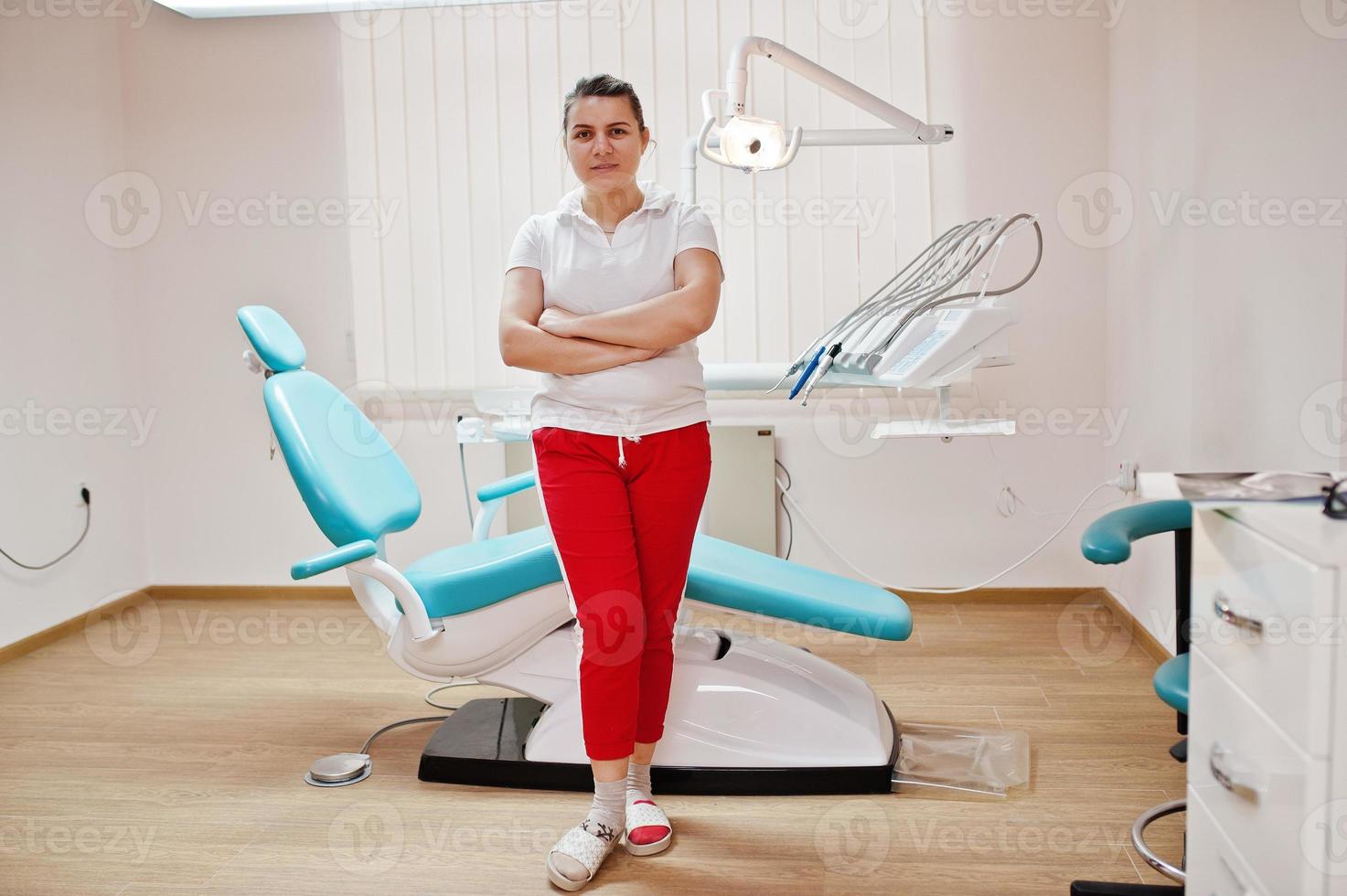 porträtt av kvinnlig tandläkare kvinna korsade armarna står i hennes tandläkare kontor nära stol. foto