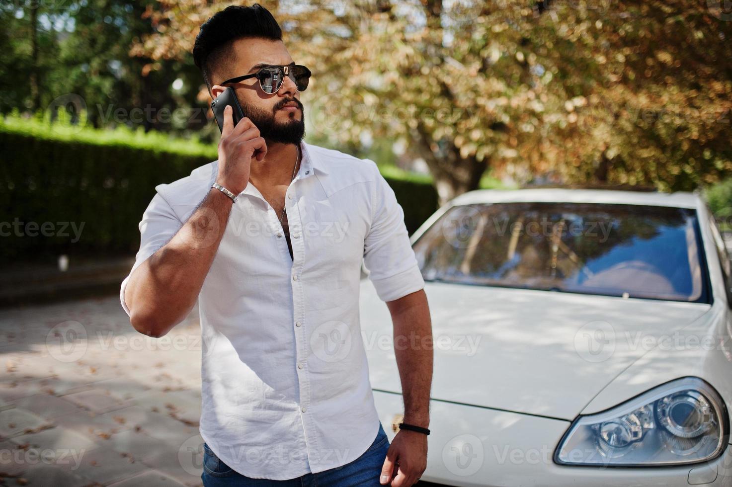 snygg lång arabisk man modell i vit skjorta, jeans och solglasögon poserade på gatan i staden. skägg rik attraktiv arabisk kille mot vit suv bil, talar i mobiltelefon. foto