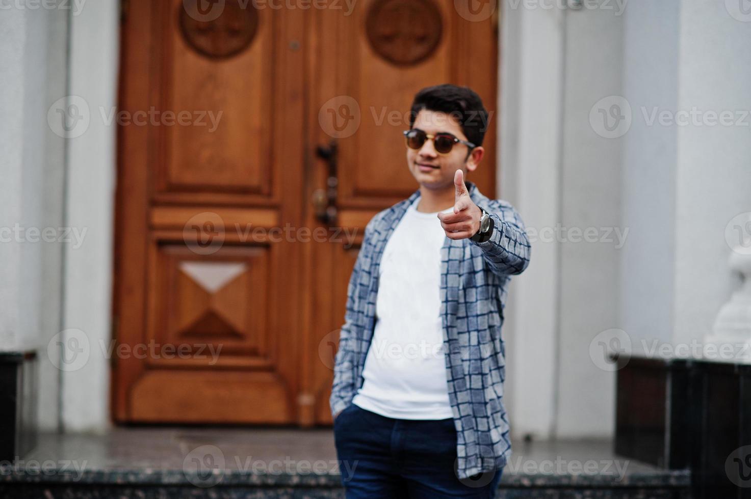 elegant indisk ung man på solglasögon bär casual poserade utomhus mot dörren till byggnaden, visa finger på kameran. foto