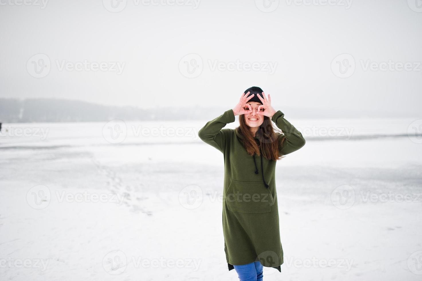 rolig flicka bär på lång grön tröja, jeans och svarta huvudbonader, vid frusen sjö i vinterdag. foto