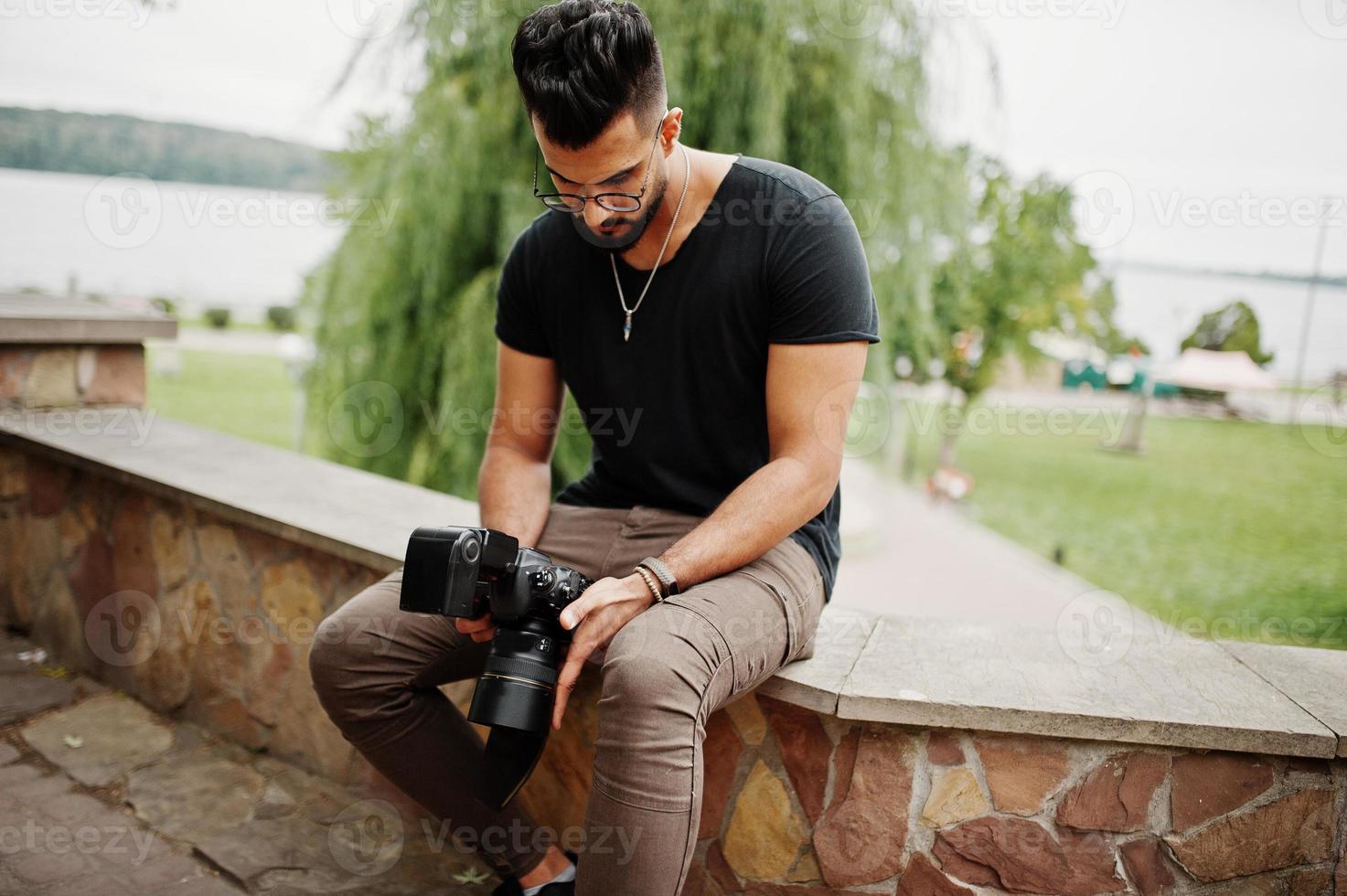 grymt vackert högt arabiskt skägg macho man fotograf i glasögon och svart t-shirt med professionell kamera till hands. foto