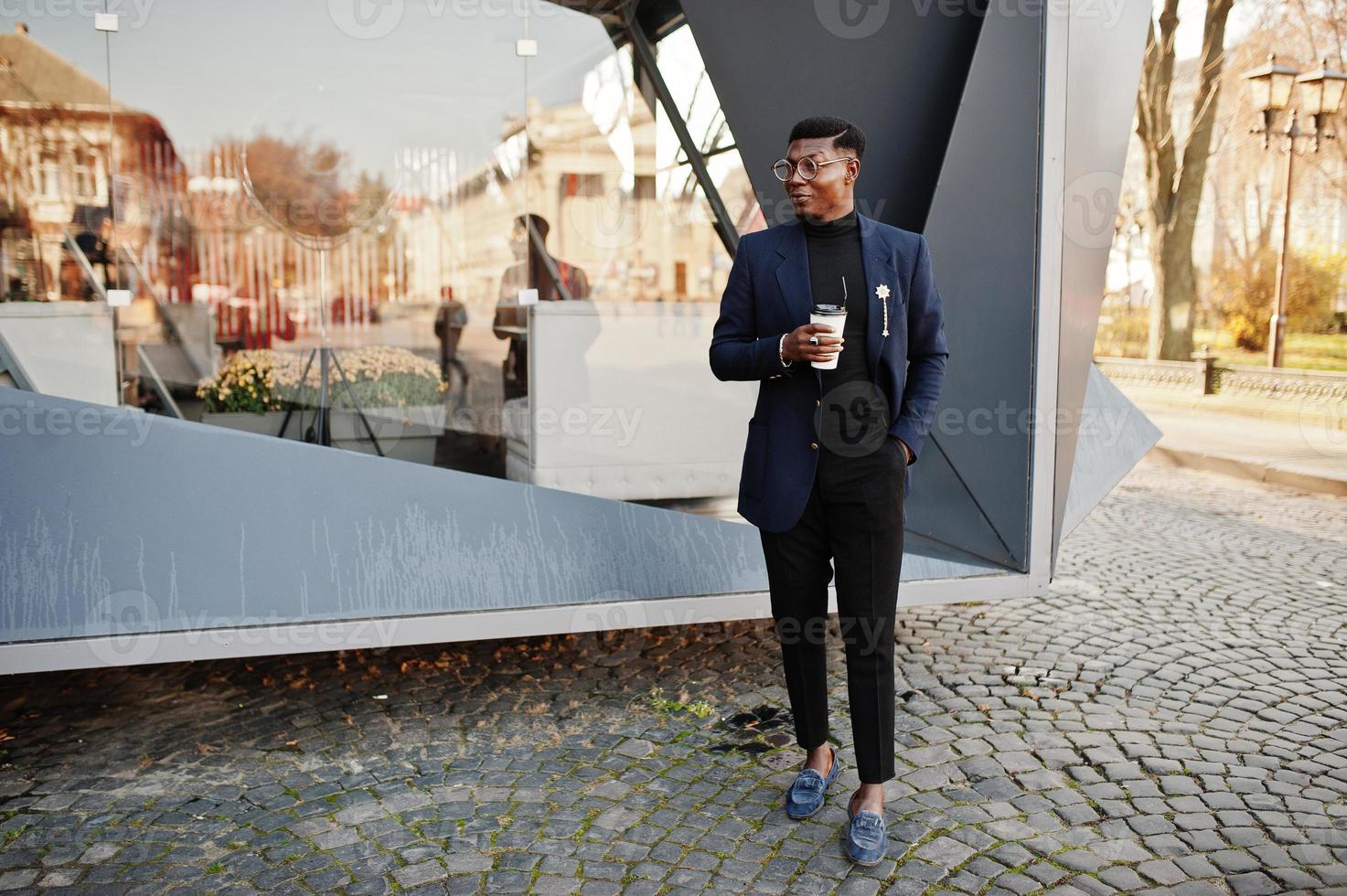 otroligt snygg afrikansk amerikansk man bär på blå kavaj med brosch, svart polotröja och glasögon poserade på gatan. fashionabla svart kille med kopp kaffe. foto