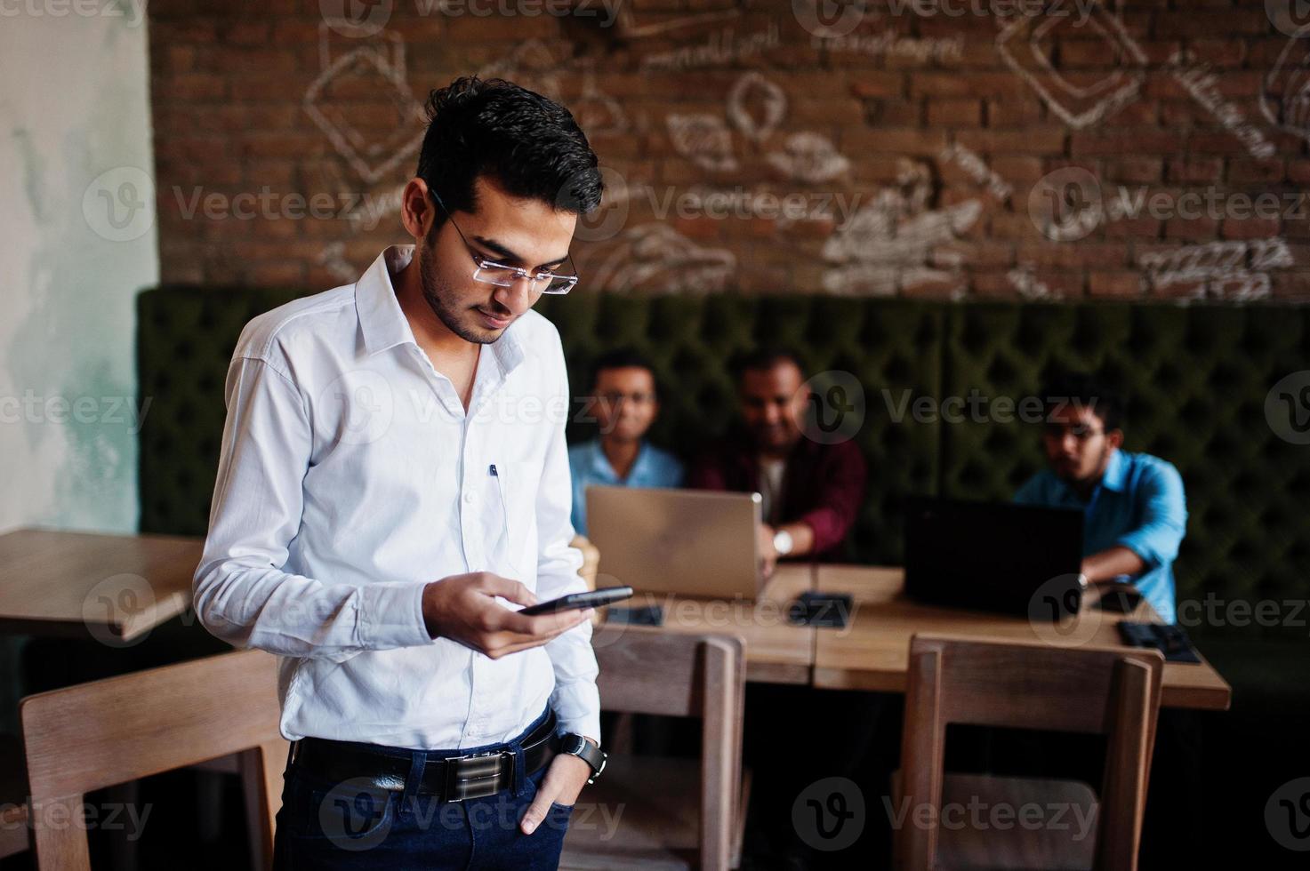grupp av fyra sydasiatiska män poserade på affärsmöte i café. indianer arbetar med bärbara datorer tillsammans med olika prylar och pratar. indisk man med mobiltelefon. foto