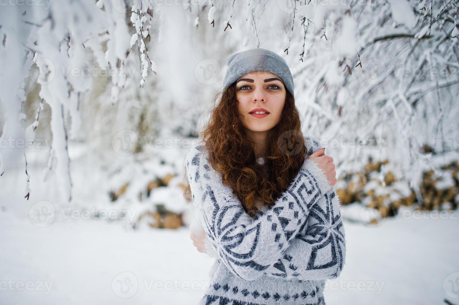 söt lockig tjej i tröja och huvudbonader på snöig skogspark på vintern. foto