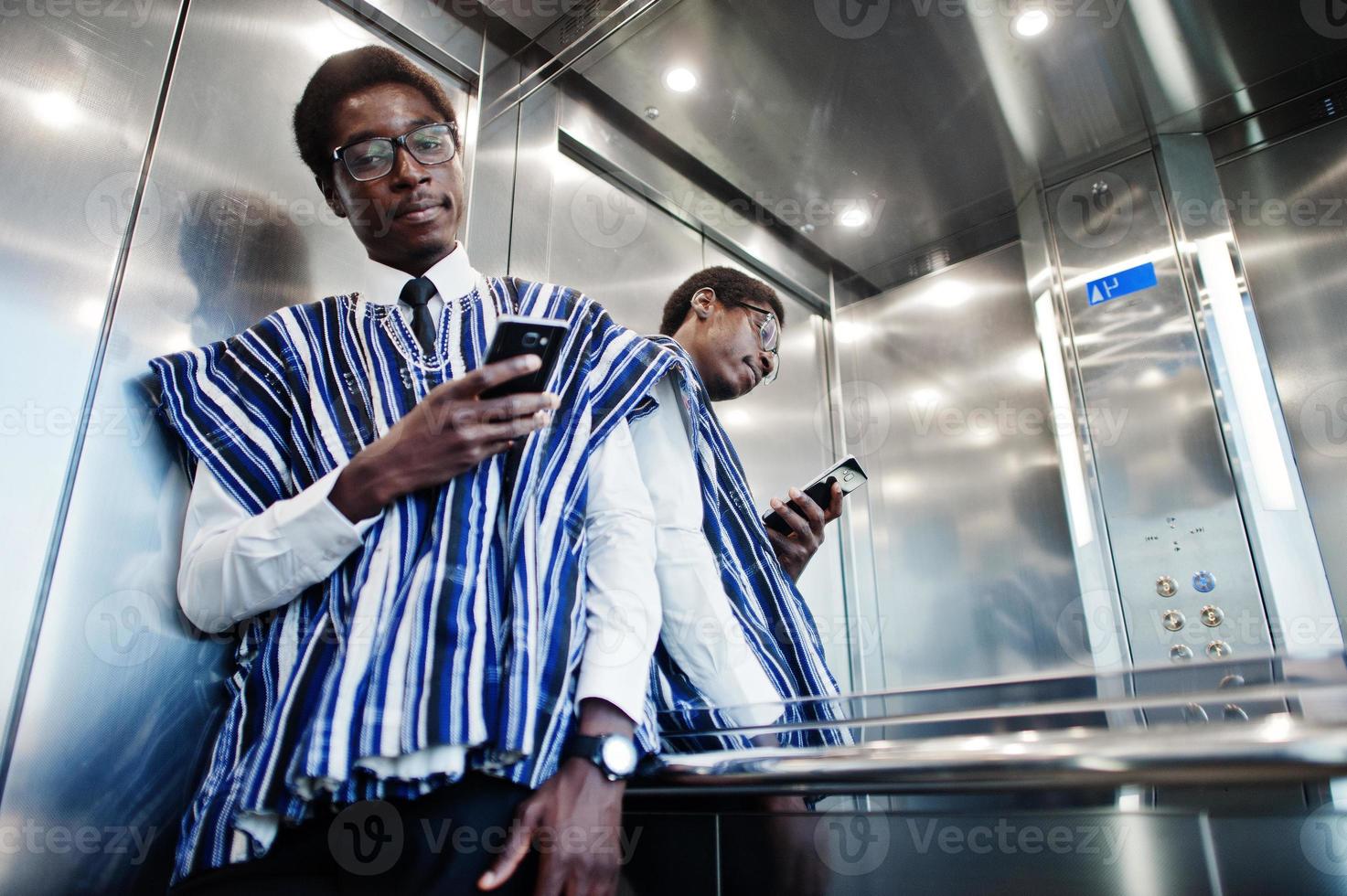 afrikansk man i traditionella kläder och glasögon med mobiltelefon vid hissen eller modern hiss. foto