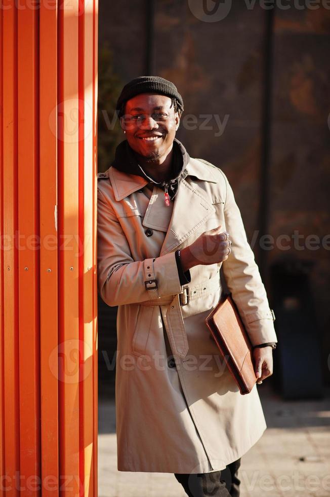 stilig afrikansk amerikansk man poserar utanför i svart hatt och beige kappa med mapp i handen. foto