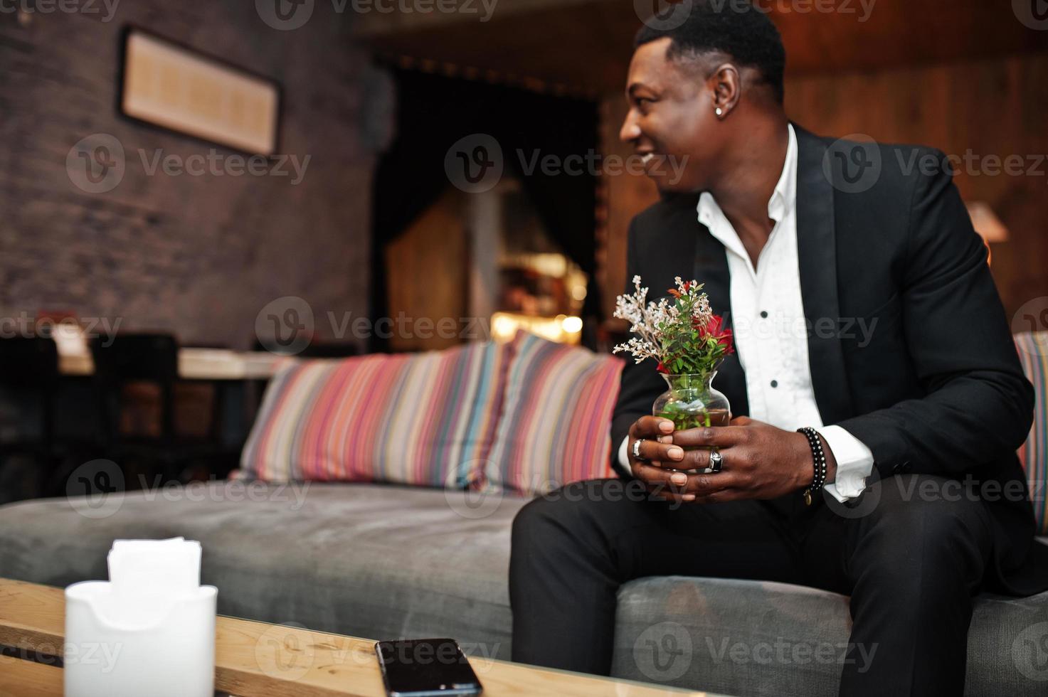 stark kraftfull afroamerikansk man i svart kostym sitter på soffan och håller vas med blommor inomhus relaxrum. foto