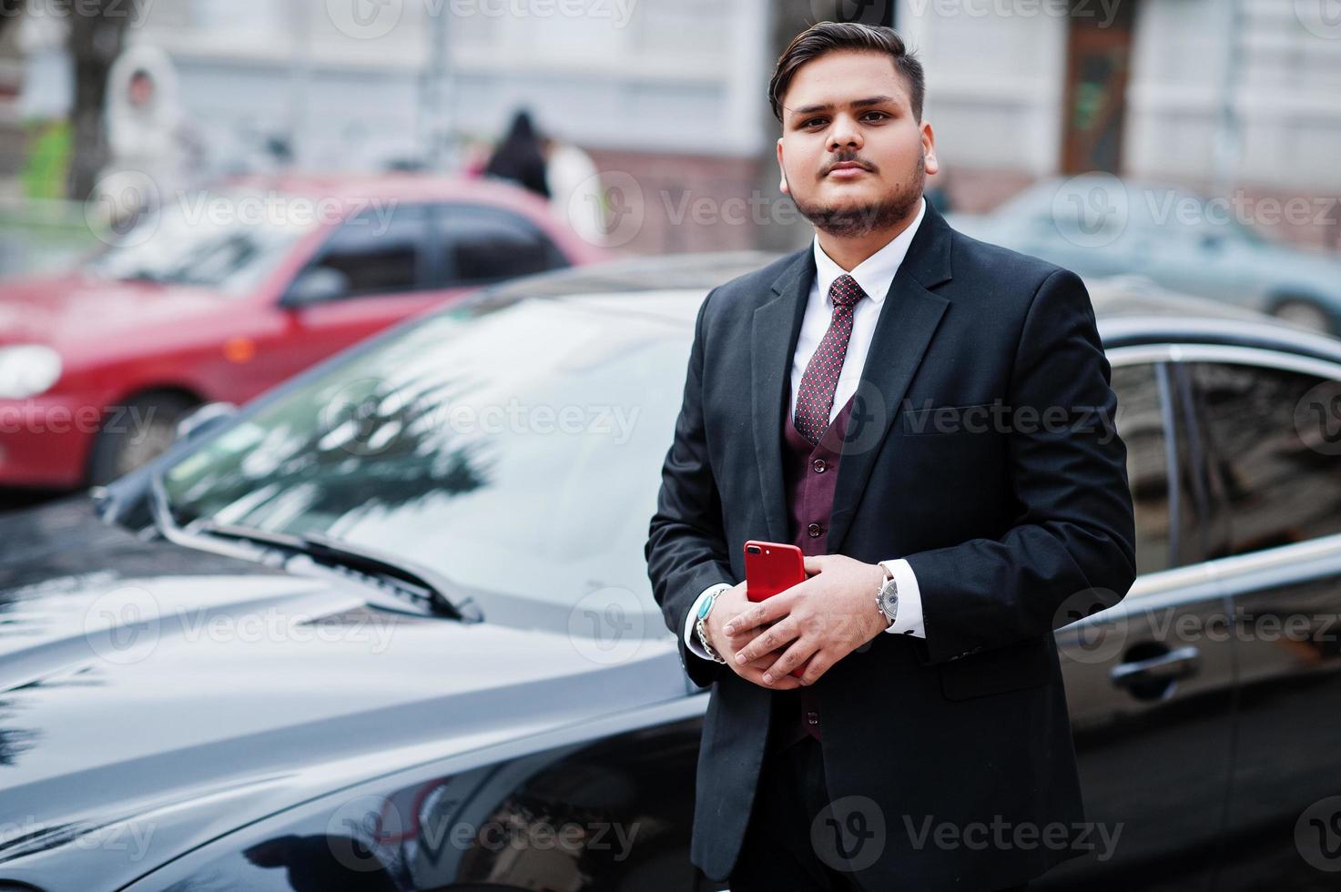 snygg indisk affärsman i högtidskläder med mobiltelefon stående mot svart affärsbil på gatan i staden. foto