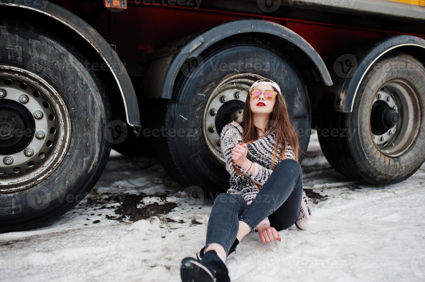 brunett snygg casual tjej i mössa och solglasögon sitter mot lastbil hjul. foto