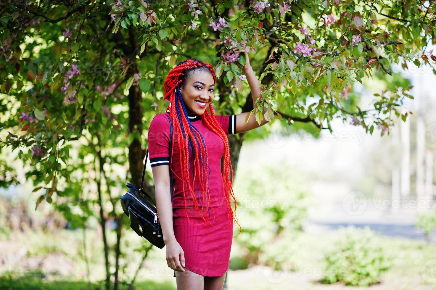 söt och smal afrikansk amerikansk tjej i röd klänning med dreadlocks poserade utomhus i vårparken. stilren svart modell. foto