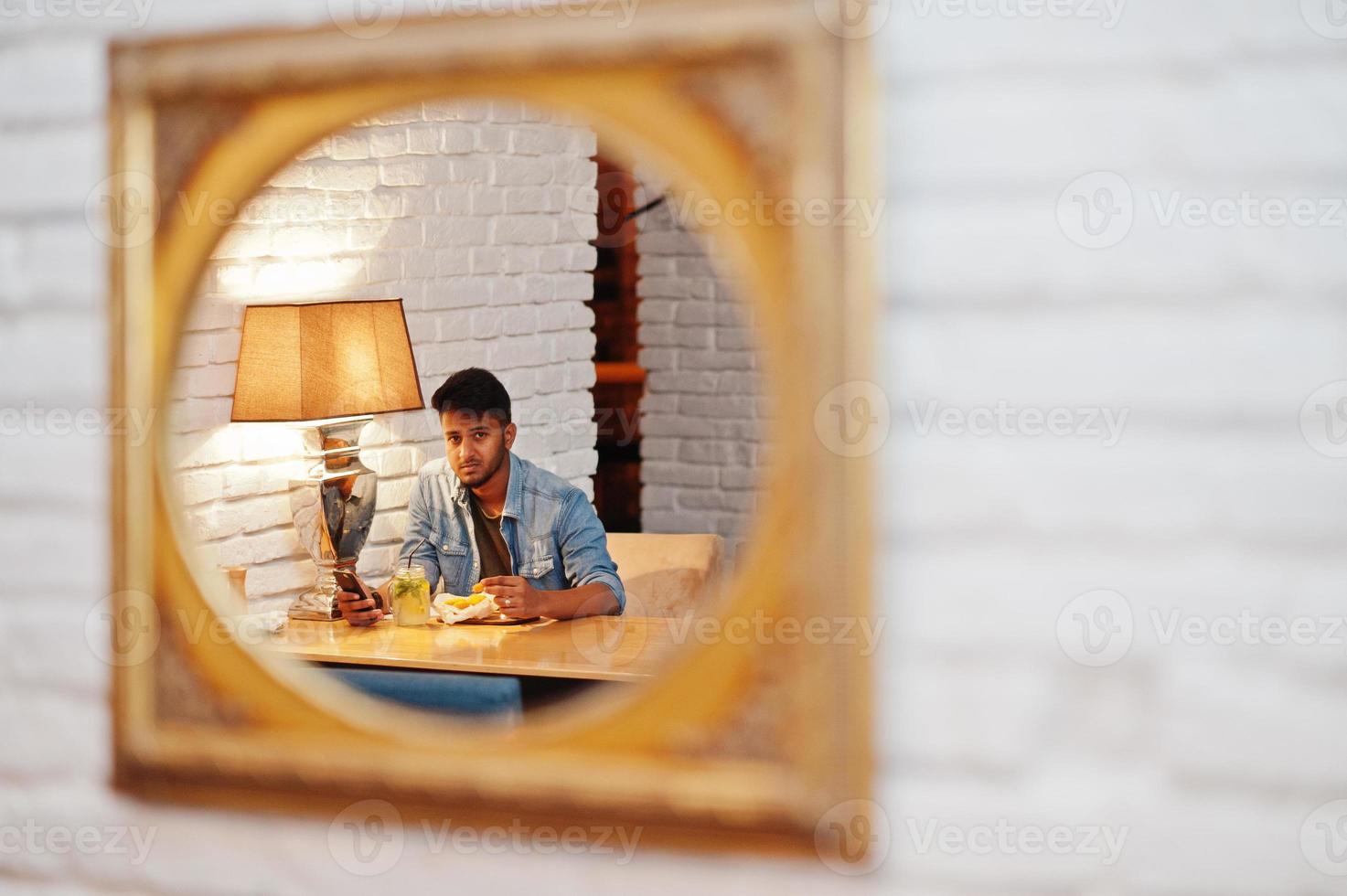 porträtt av stilig framgångsrik skäggig sydasiatisk, ung indisk frilansare i blå jeansskjorta som sitter på café med kycklingnuggets och lemonad. foto