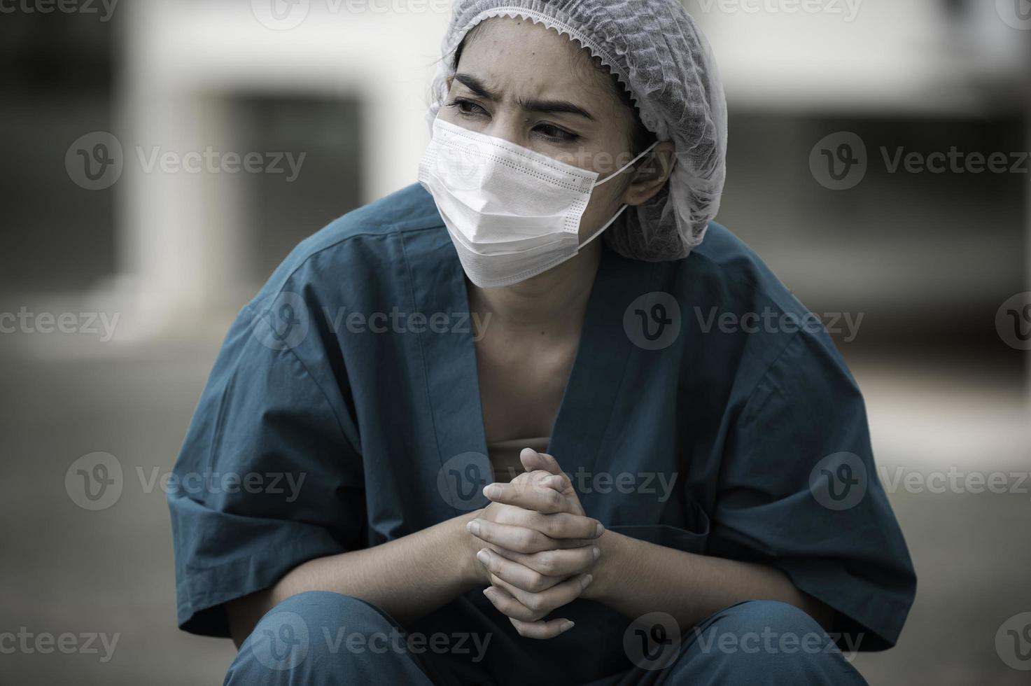 trött deprimerad kvinnlig asiatisk skrubbsköterska bär ansiktsmask blå uniform sitter på sjukhusgolvet, ung kvinna läkare stressad av hårt arbete foto