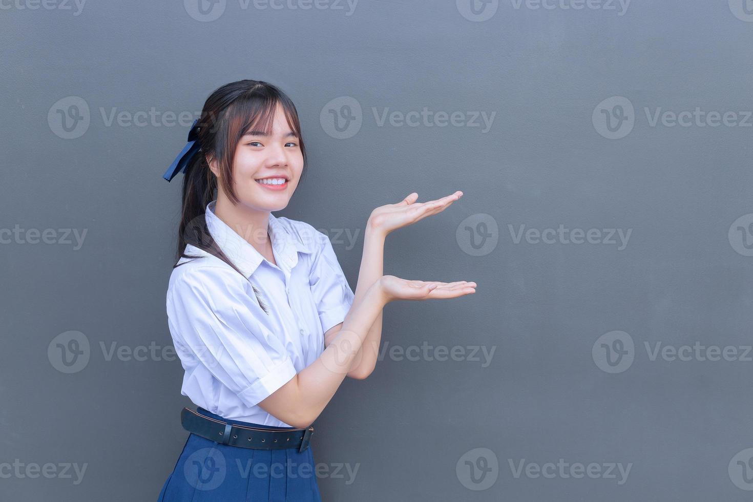 söt asiatisk gymnasieelev flicka i skoluniformen med leenden självsäkert medan hon tittar på kameran för att presentera något glatt med grått i bakgrunden. foto