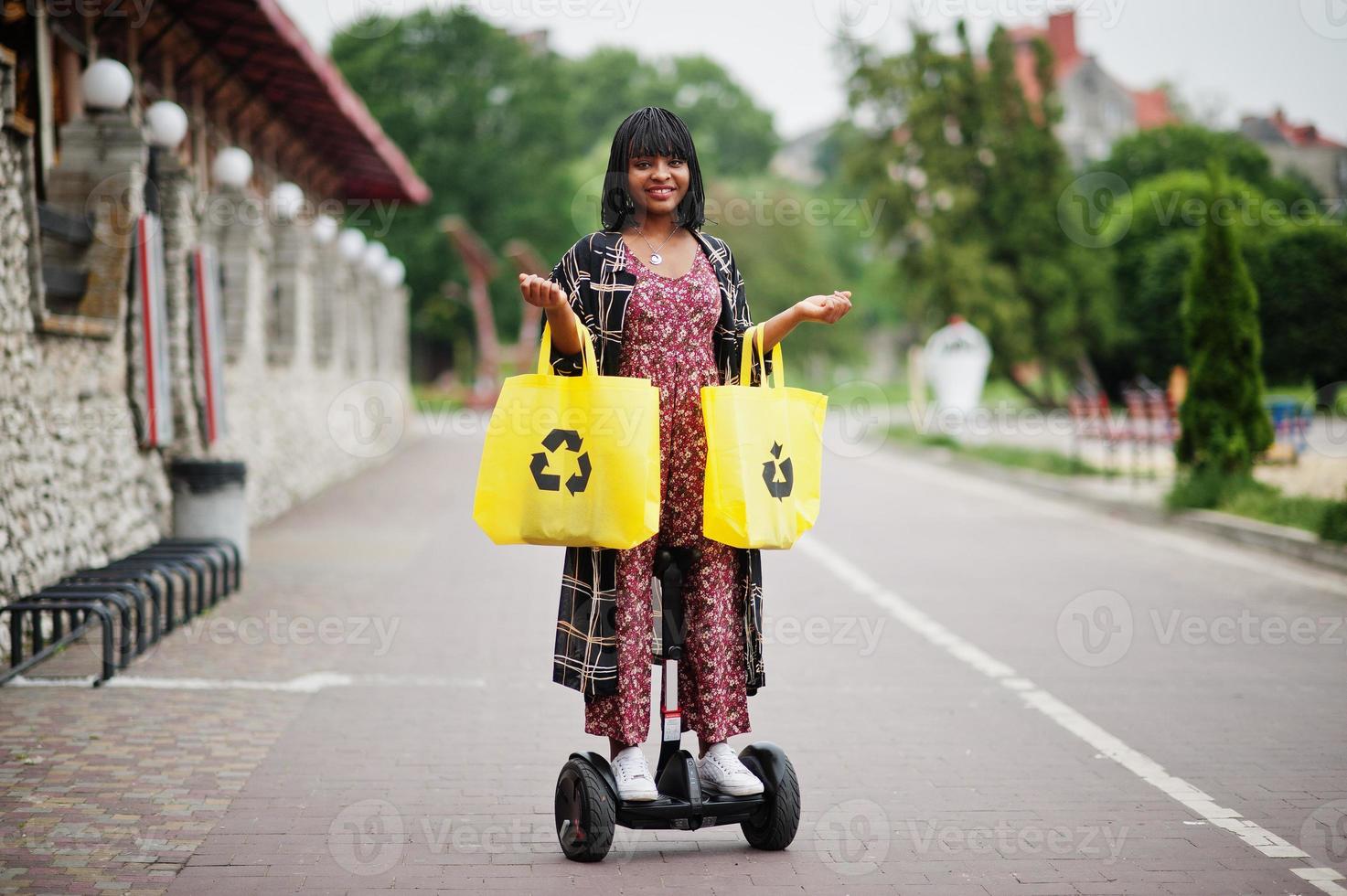 vacker afrikansk amerikansk kvinna som använder segway eller hoverboard. svart flicka med gul duk eco väskor återvinning symbol. foto