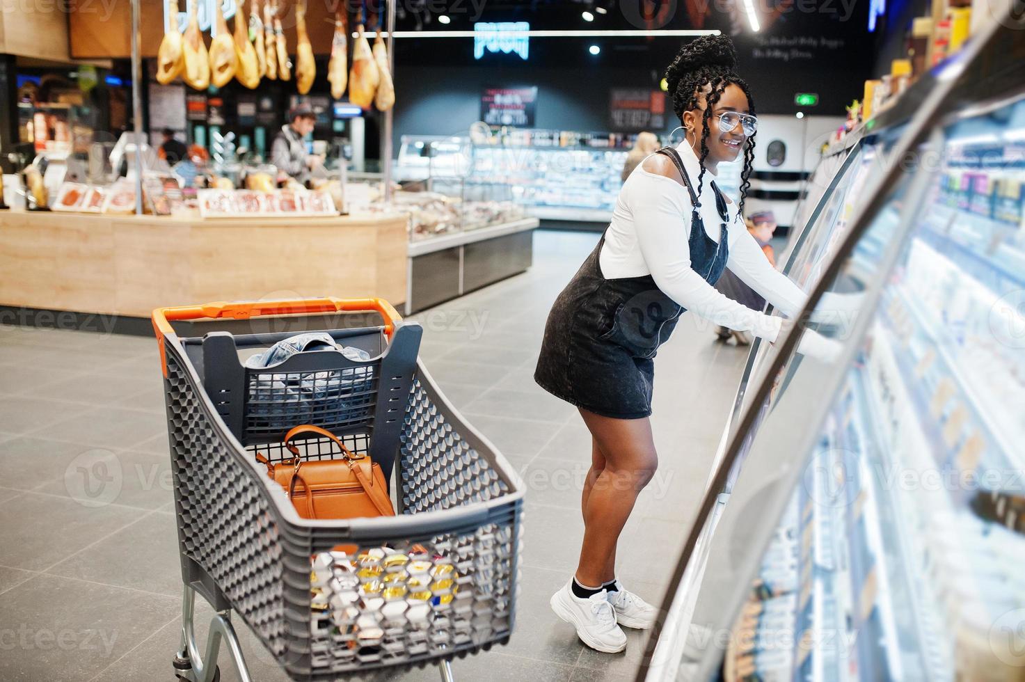 afrikansk kvinna med kundvagn väljer yoghurtflaska från kylskåpet på snabbköpet. foto