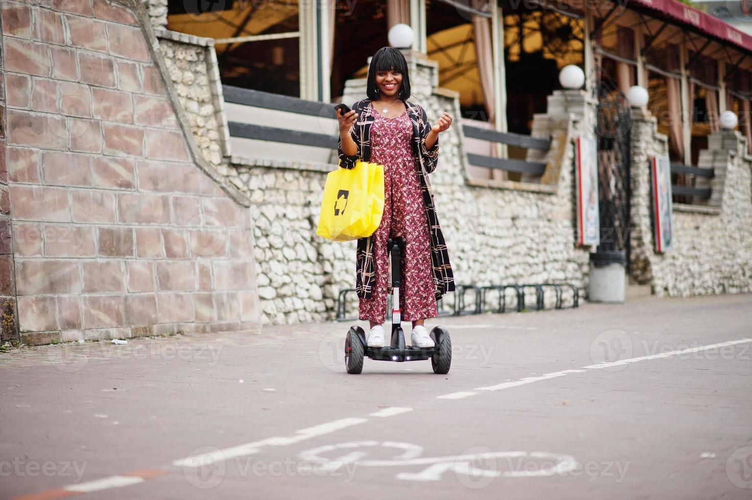 vacker afrikansk amerikansk kvinna som använder segway eller hoverboard. svart flicka med gul duk eco väskor återvinning symbol. foto