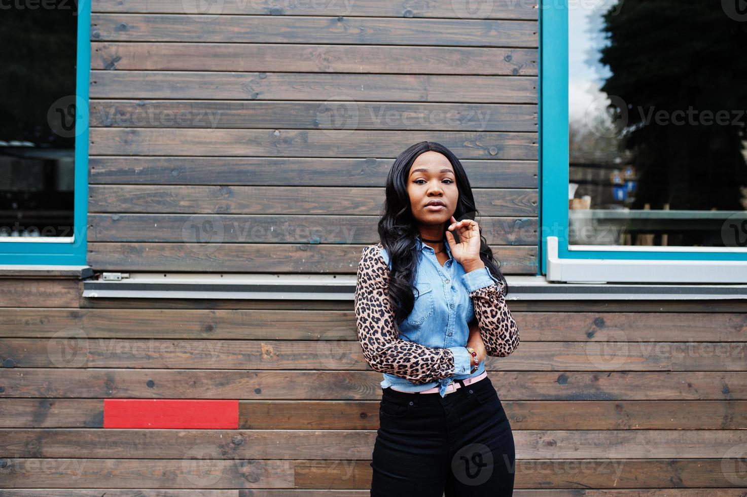 hipster afrikansk amerikansk flicka klädd i jeansskjorta med leopardärmar poserar på gatan mot trähus med fönster. foto