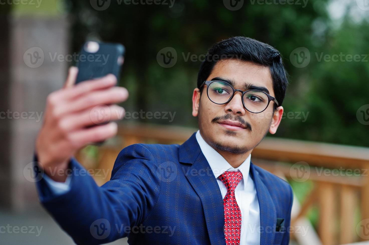 indisk ung man vid glasögon, bär kostym med röd slips poserad utomhus och gör selfie på mobiltelefon. foto