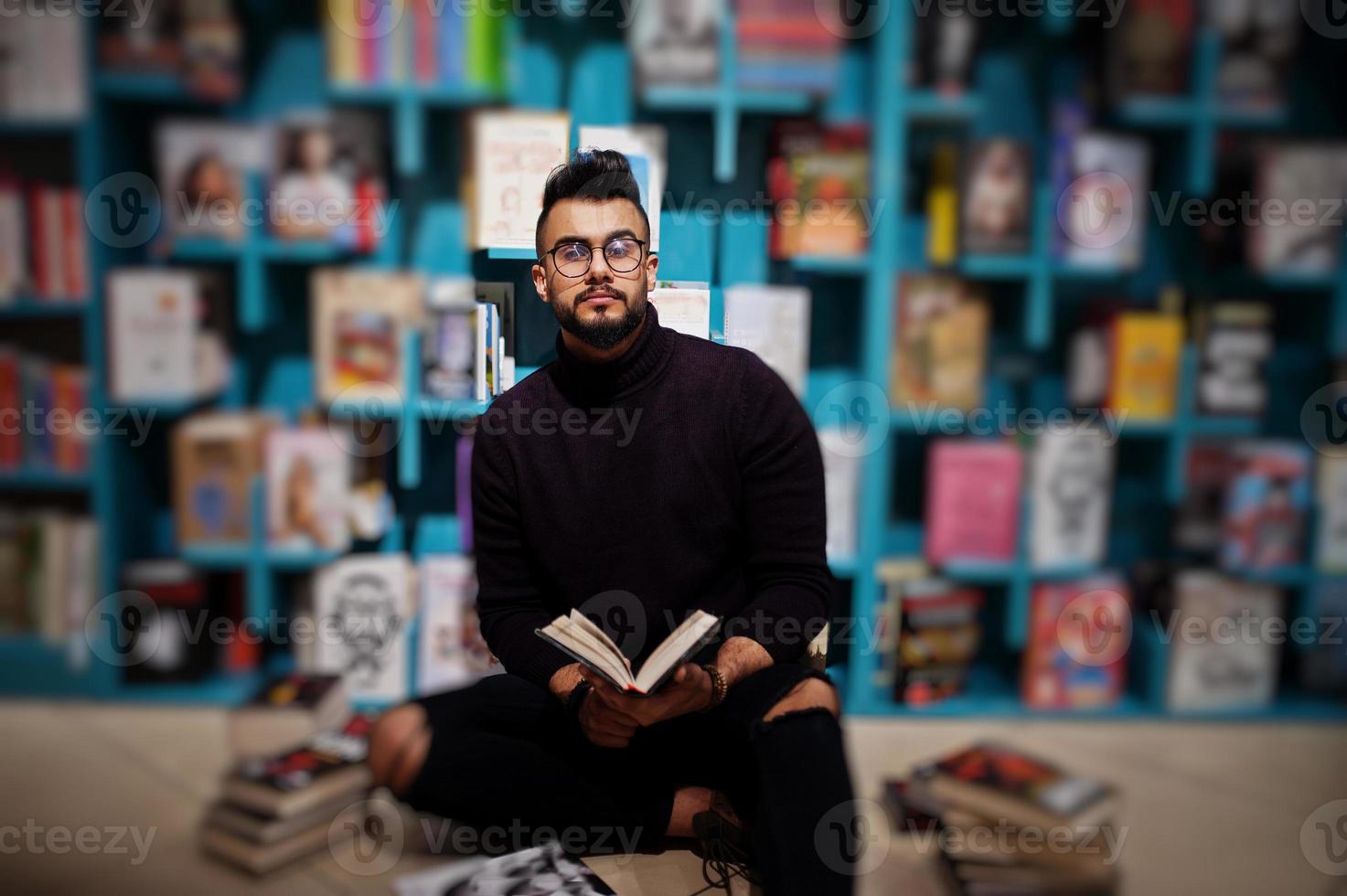 lång smart arab student man, bära på violett turtleneck och glasögon, på biblioteket sitter mot bokhyllor. foto