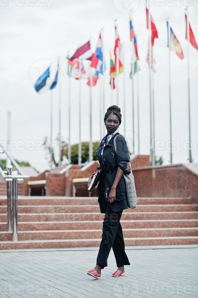 afrikansk student kvinna poserade med ryggsäck och skolsaker på gården till universitetet, mot flaggor från olika länder. foto
