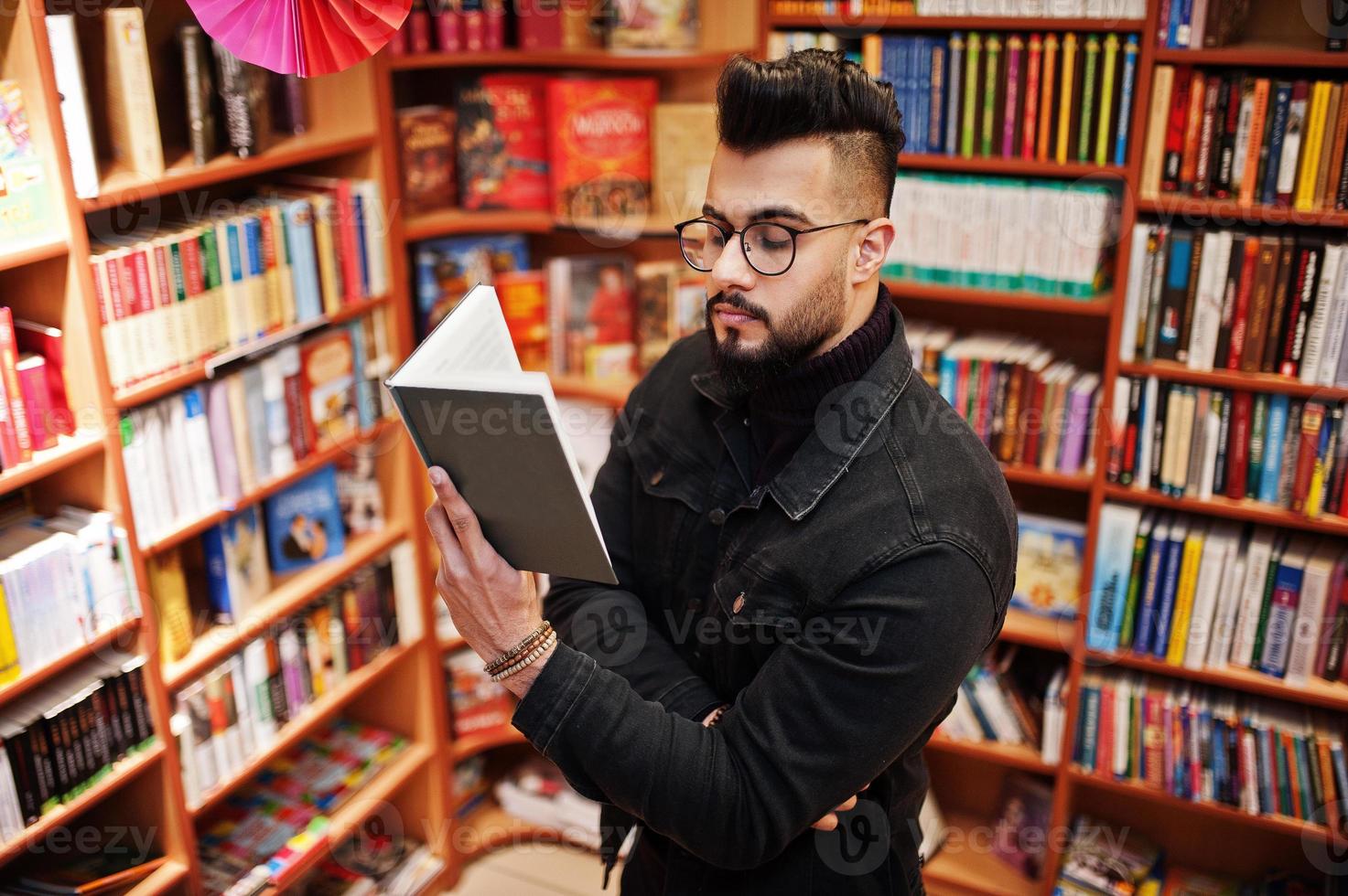 lång smart arabisk studentman, bär på svart jeansjacka och glasögon, på biblioteket med bok vid händerna. foto