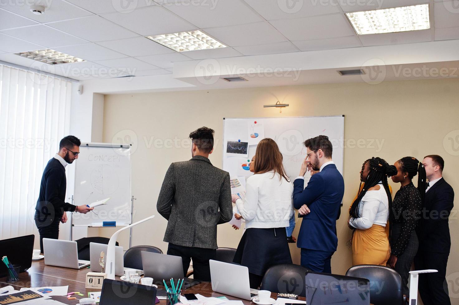 affärscoach för blandad ras som presenterar rapporten som står nära whiteboardtavlan och pekar på försäljningsstatistik som visas på diagram och diagram lär olika företagsmedlemmar samlade i konferensrummet. foto