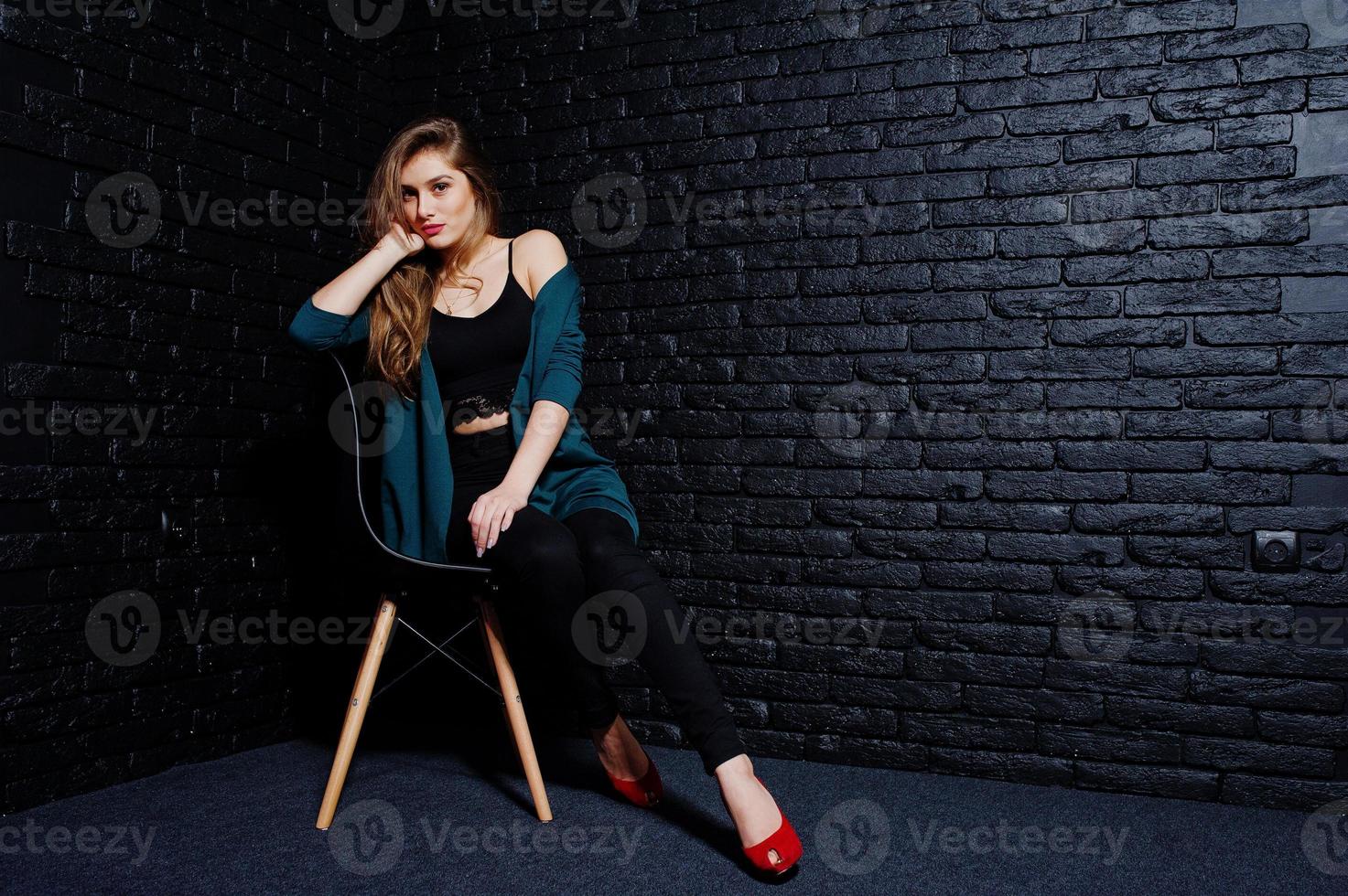 stilig brunett tjej bär på svart och grön jacka i röda höga klackar, sitter och poserar på stol i studio mot mörk tegelvägg. studio modell porträtt. foto