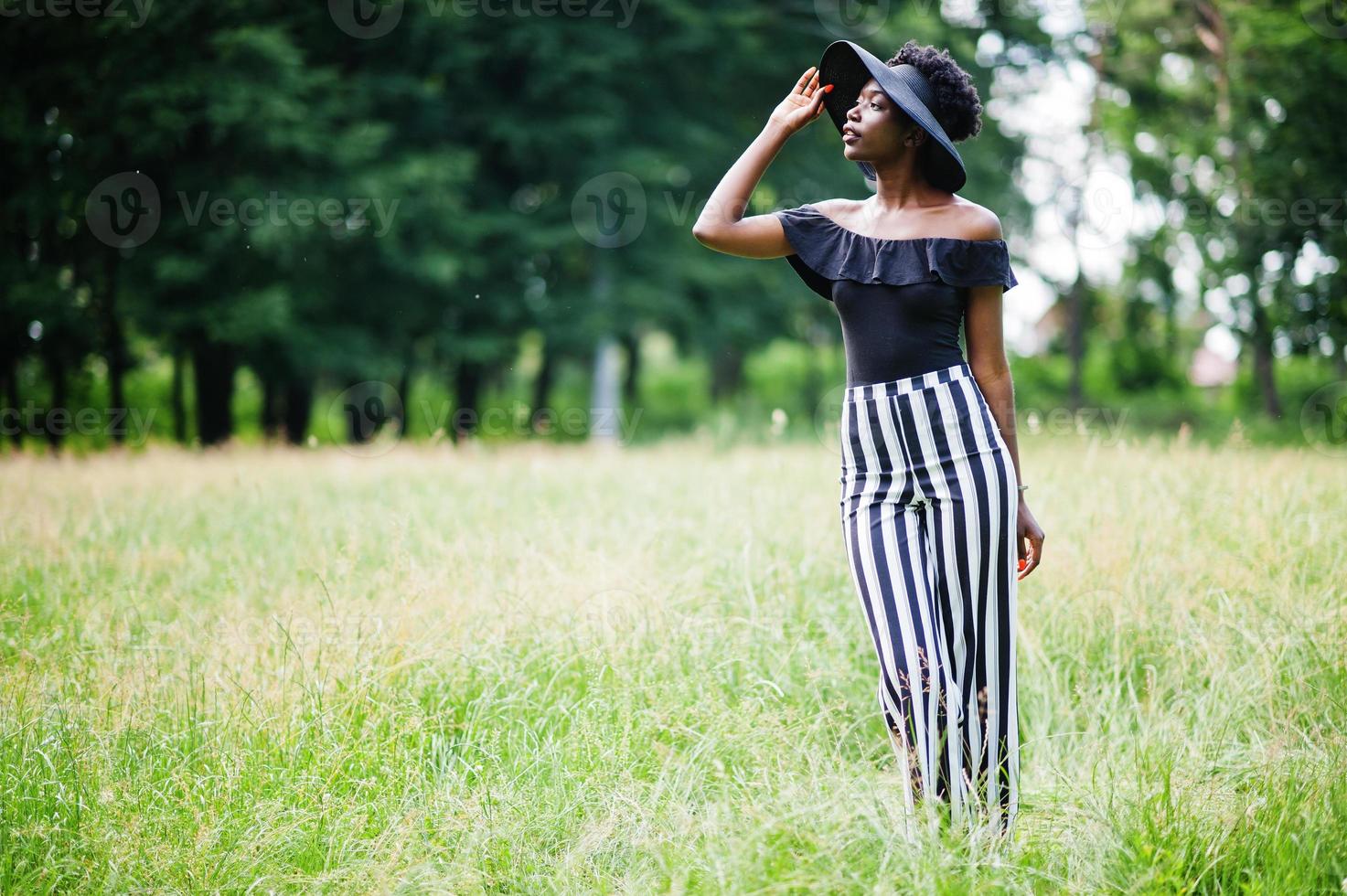 porträtt av underbar afrikansk amerikansk kvinna 20-talet i slitage i svarta och vita randiga byxor och sommarhatt poserar på grönt gräs i parken. foto