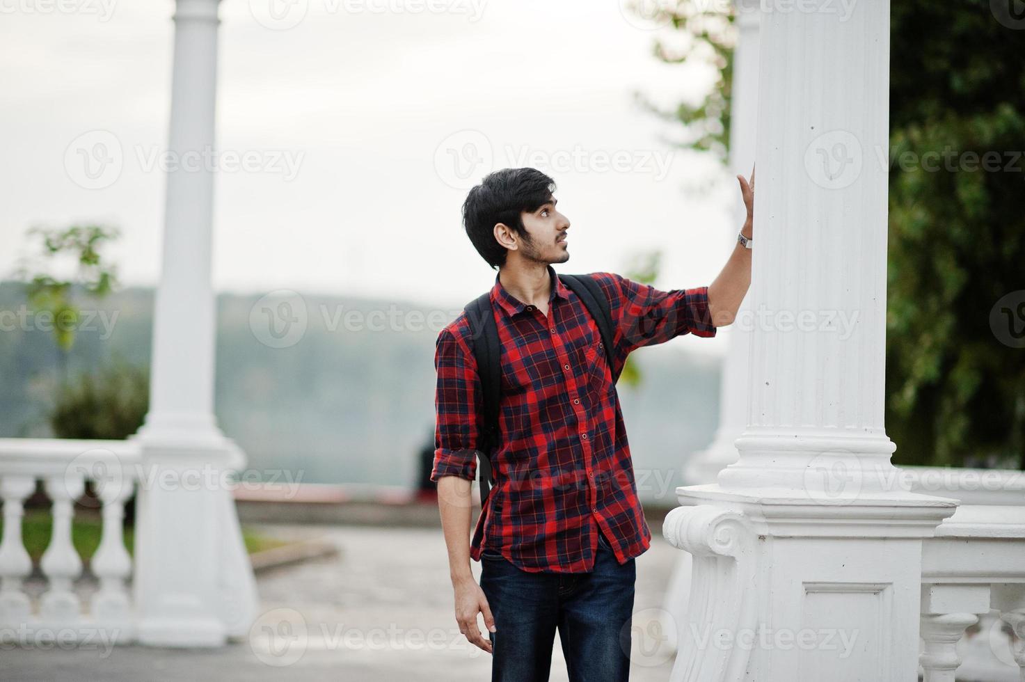 ung indisk student man på rutig skjorta och jeans med ryggsäck poserade på kvällen stad mot kolumn. foto