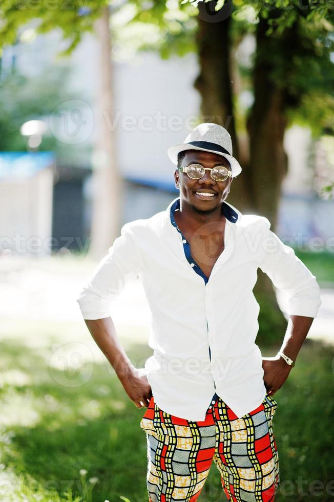 snygg afrikansk amerikansk man i vit skjorta och färgade byxor med hatt och glasögon poserade utomhus. svart fashionabla modellpojke. foto