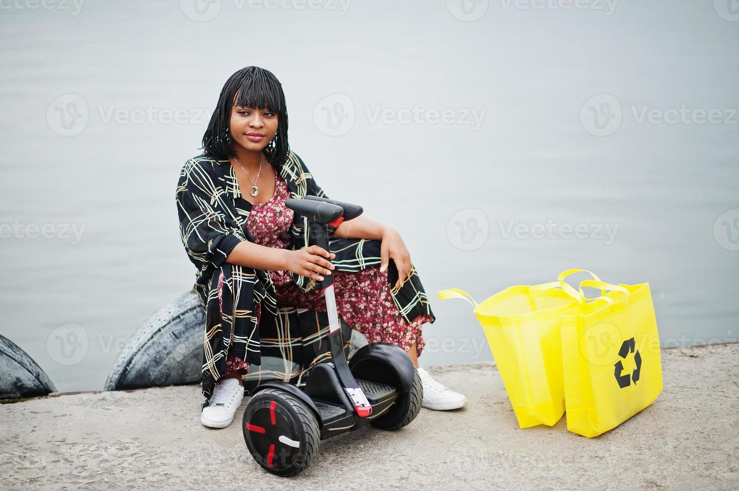 vacker afrikansk amerikansk kvinna sitter nära segway eller hoverboard. svart flicka med gul duk eco väskor återvinning symbol. foto