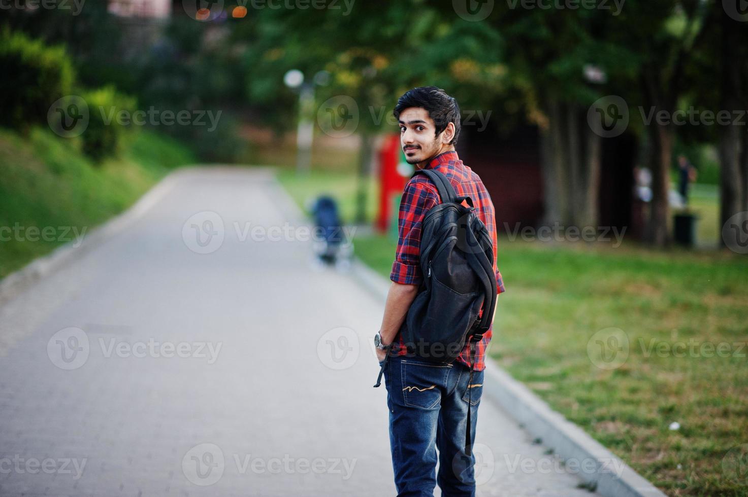 ung indisk student man på röd rutig skjorta och jeans med ryggsäck poserade på gatan. foto