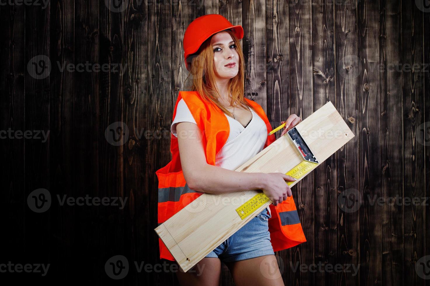 ingenjör kvinna i orange skydda hjälm och bygga jacka mot trä bakgrund håller styrelse och linjal. foto