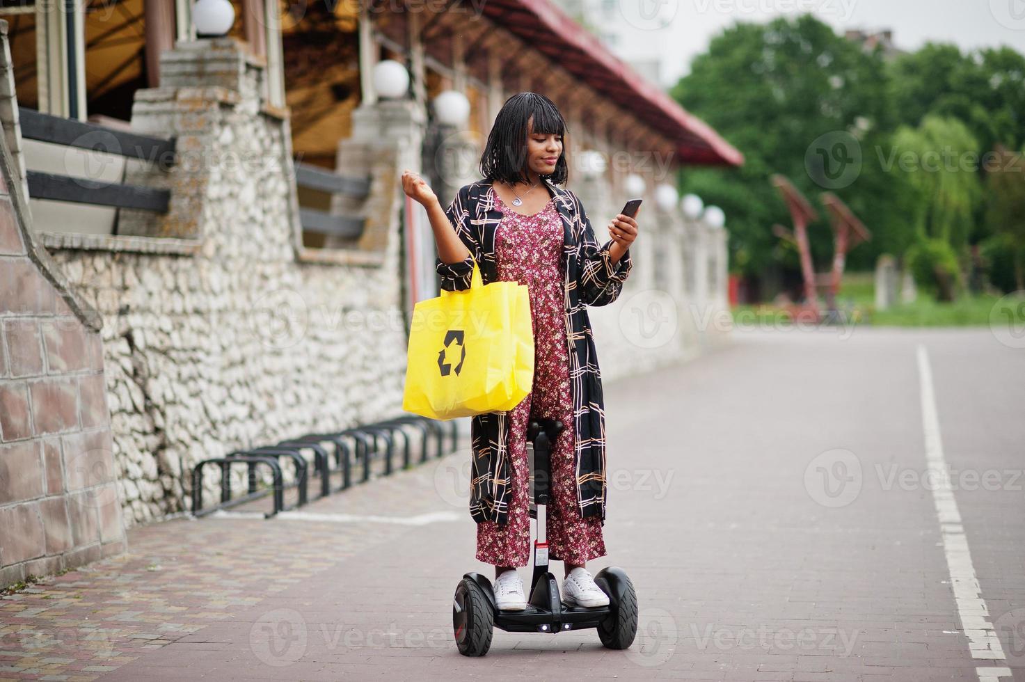 vacker afrikansk amerikansk kvinna som använder segway eller hoverboard och mobiltelefon. svart flicka med gul duk eco väskor återvinning symbol. foto
