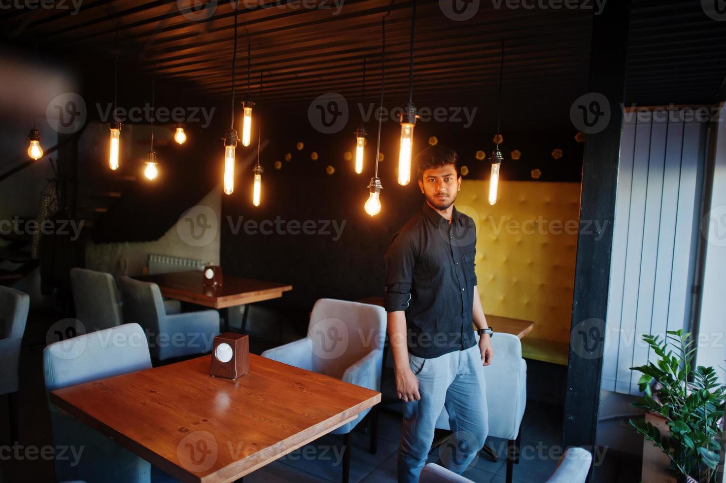 självsäker ung indisk man i svart skjorta står på café. foto