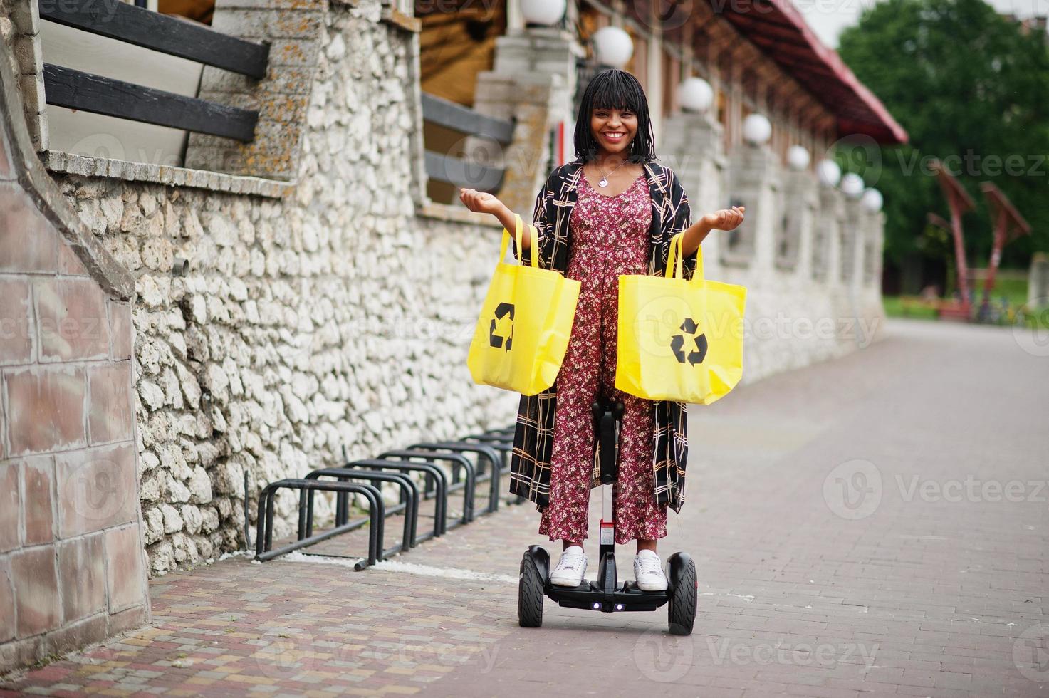 vacker afrikansk amerikansk kvinna som använder segway eller hoverboard. svart flicka med gul duk eco väskor återvinning symbol. foto