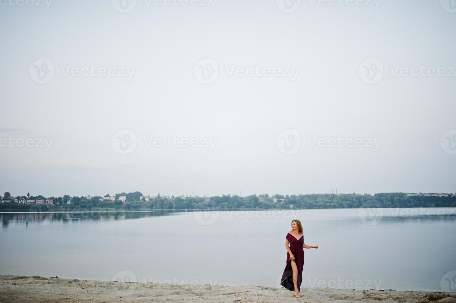 blond sensuell barfota kvinna i röd marsala klänning poserar mot sjön på sand. foto