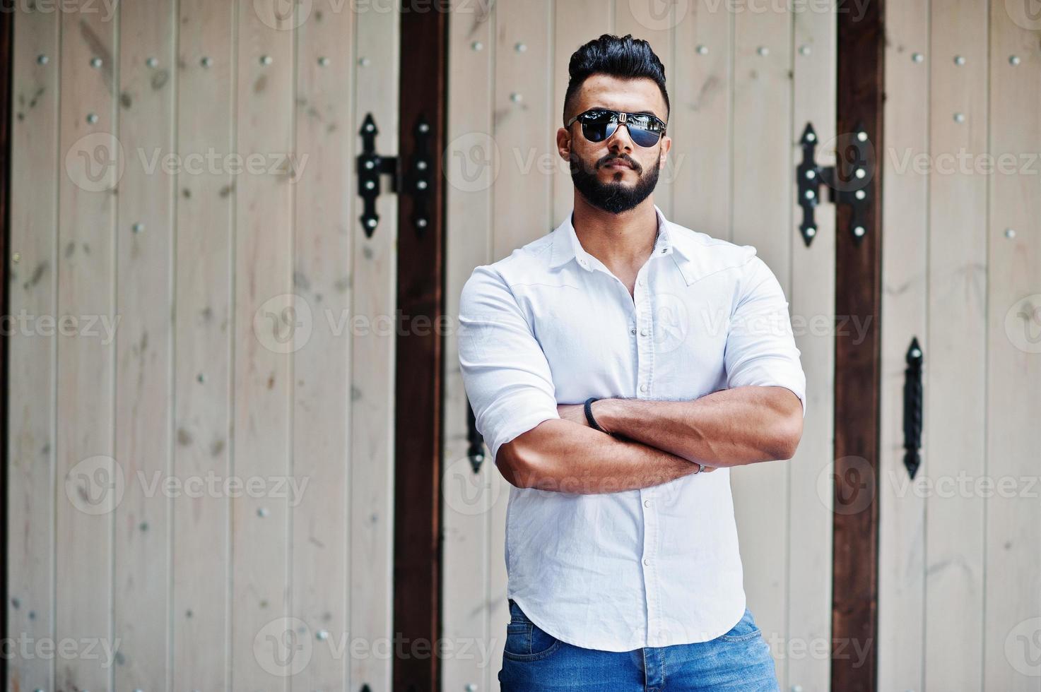 snygg lång arabisk man modell i vit skjorta, jeans och solglasögon poserade på gatan i staden. skägg attraktiv arabisk kille. foto
