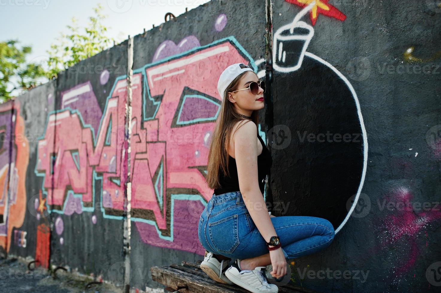 baksidan av skinkorna snygg casual hipster tjej i mössa, solglasögon och jeans slitage, lyssna musik från hörlurar av mobiltelefon mot stor graffiti vägg med stor tnt bomb. foto