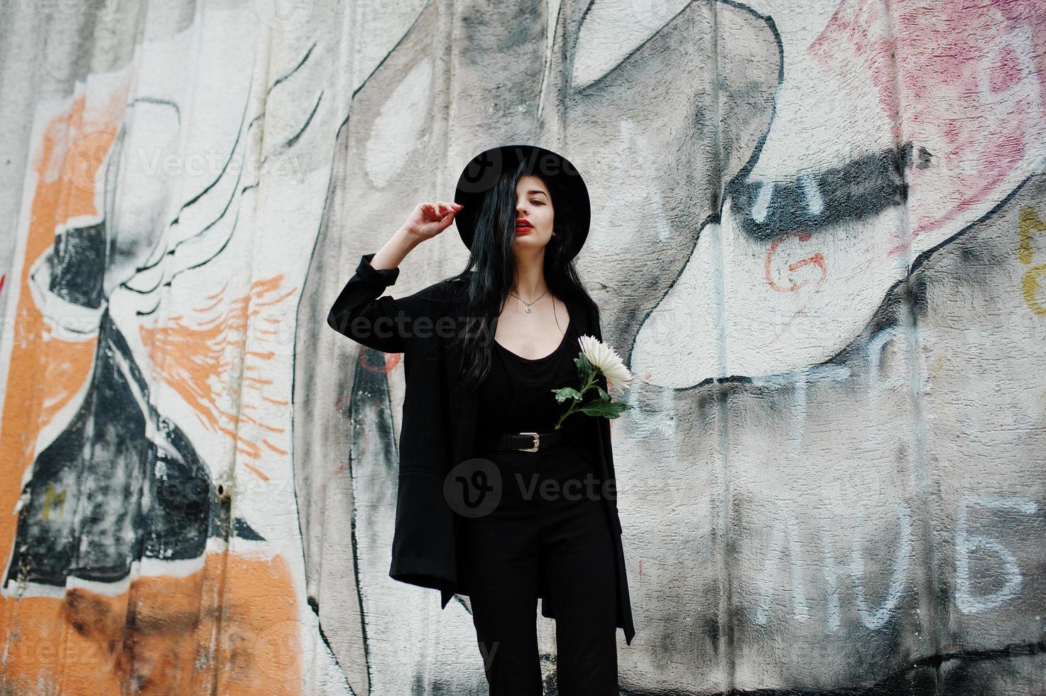 sensuell tjej helt i svarta, röda läppar och hatt. goth dramatisk kvinna håller vit krysantemum blomma mot graffiti vägg. foto