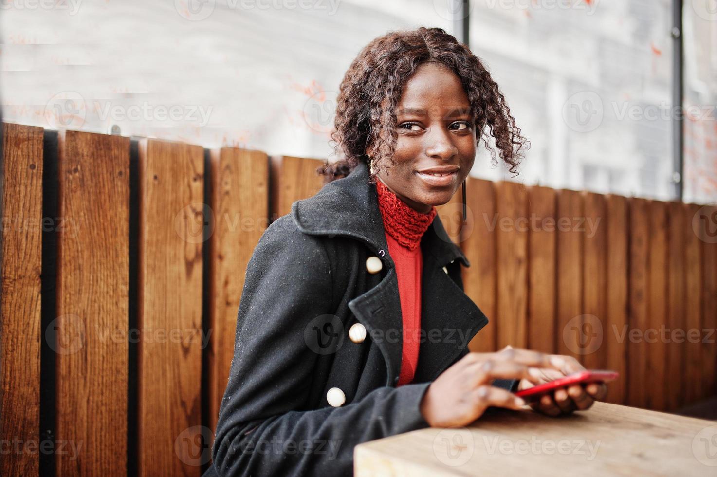 porträtt av en lockig afrikansk kvinna som bär en fashionabel svart kappa och röd polotröja sitter med mobiltelefonen vid händerna. foto