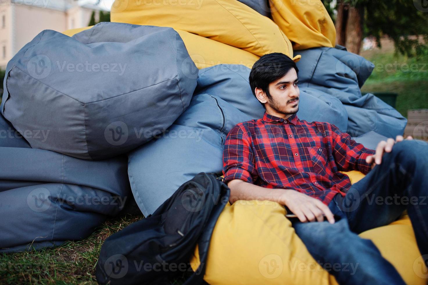 ung indisk student man på rutig skjorta och jeans sitter och koppla av vid utomhus kuddar. foto