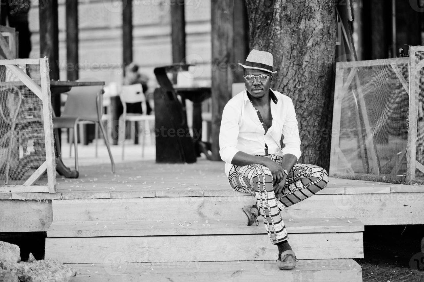 snygg afrikansk amerikansk man i vit skjorta och färgade byxor med hatt och glasögon poserade utomhus. svart fashionabla modellpojke. foto