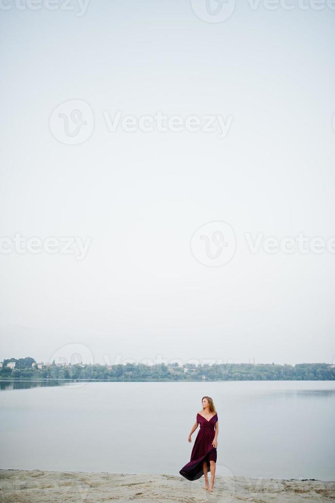 blond sensuell barfota kvinna i röd marsala klänning poserar mot sjön på sand. foto