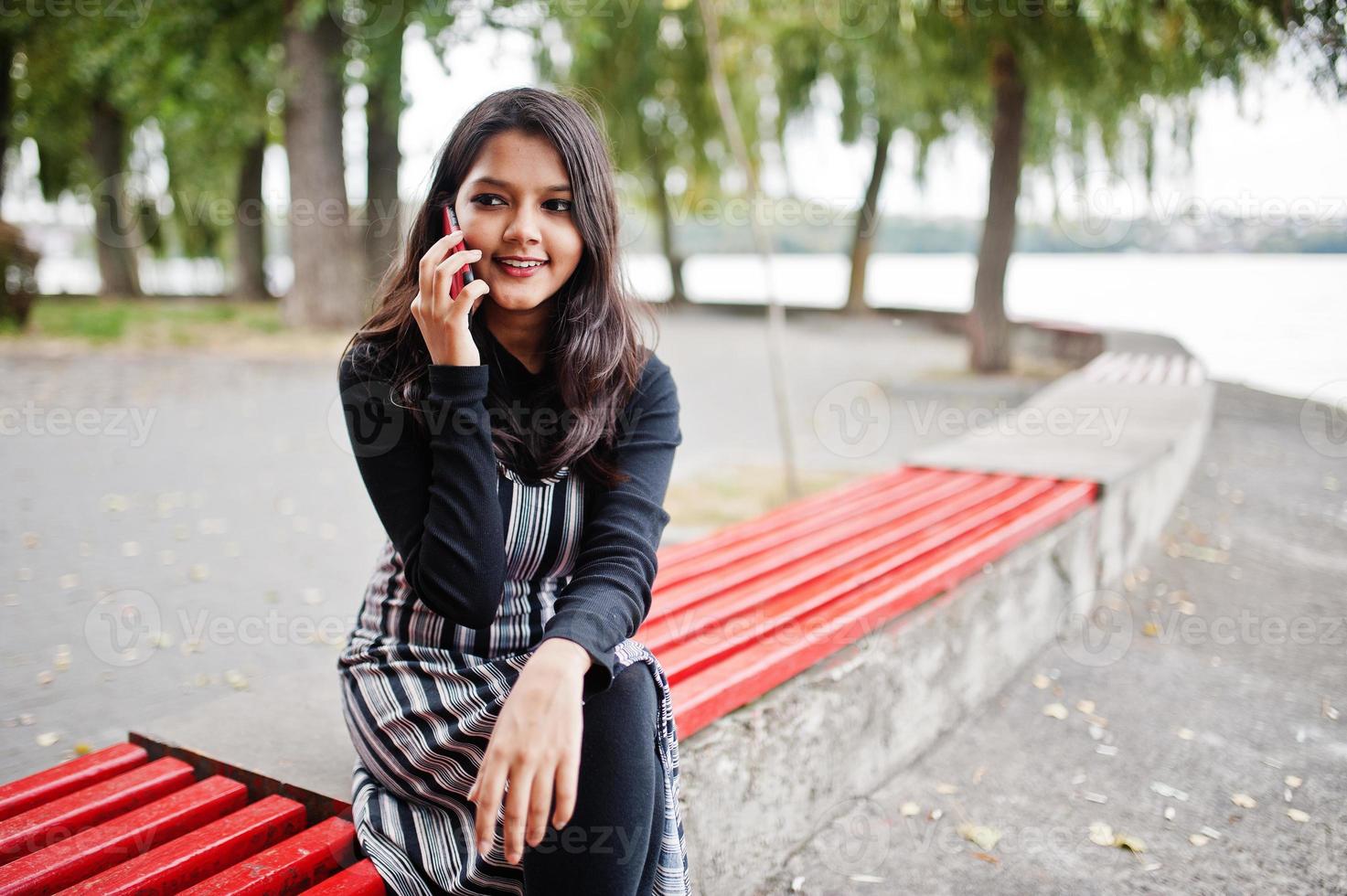 porträtt av ung vacker indisk eller sydasiatisk tonårsflicka i klänning som sitter på bänken med mobiltelefon. foto