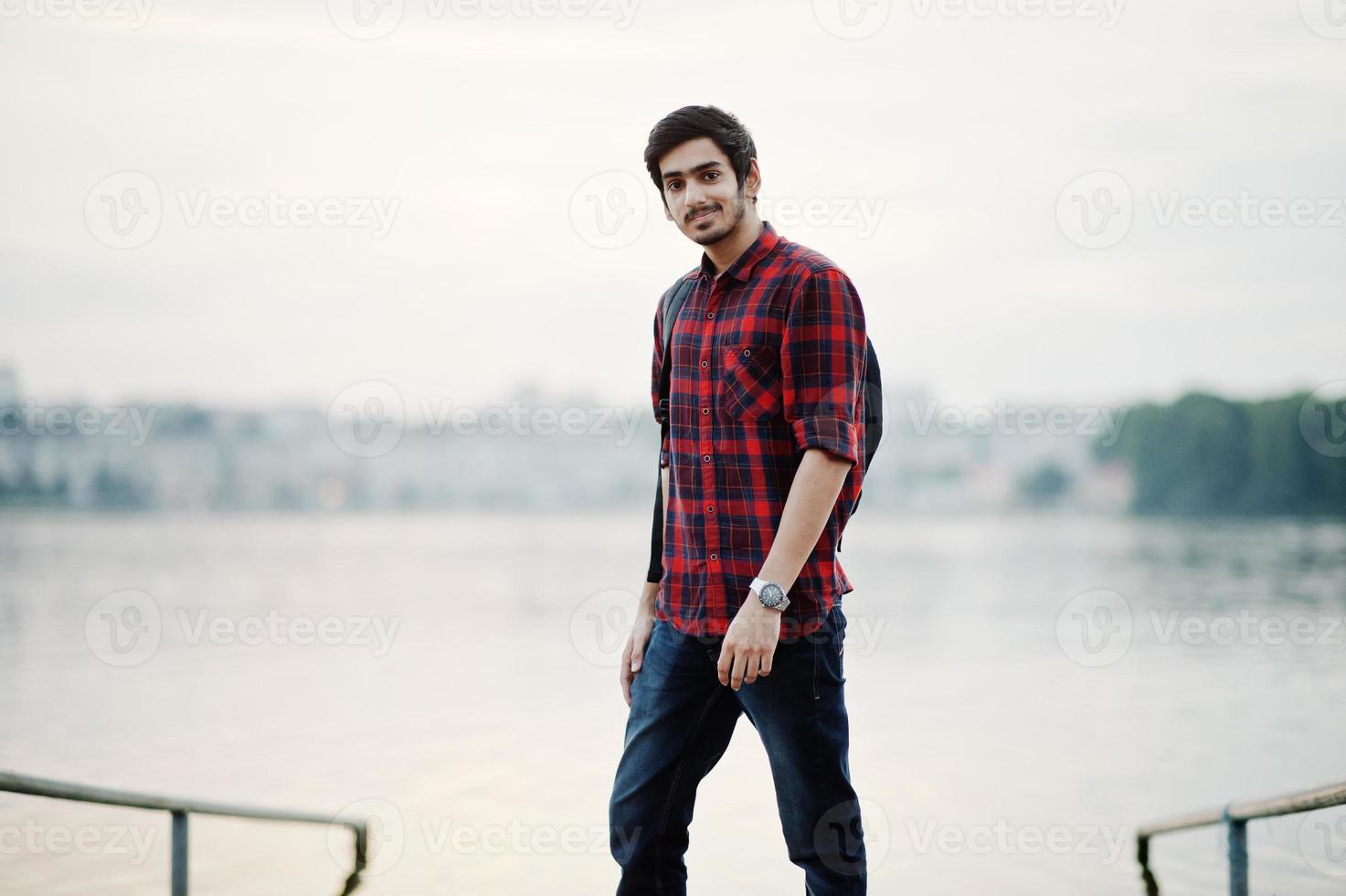 ung indisk student man på rutig skjorta och jeans med ryggsäck poserade på kvällen stad mot sjön. foto