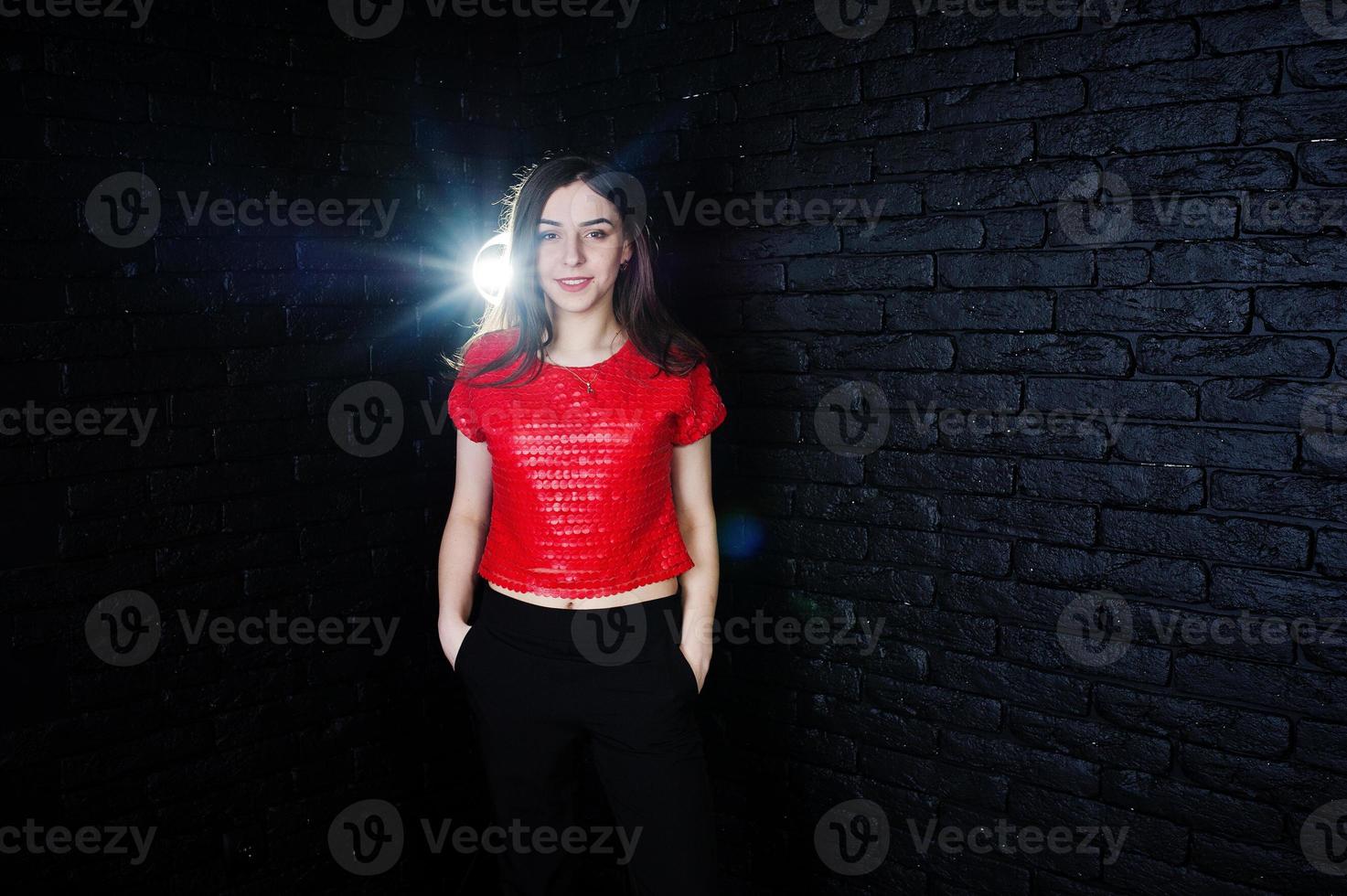 porträtt av en vacker brunett tjej i röd t-shirt och svarta byxor bredvid tegelväggen i studion. foto
