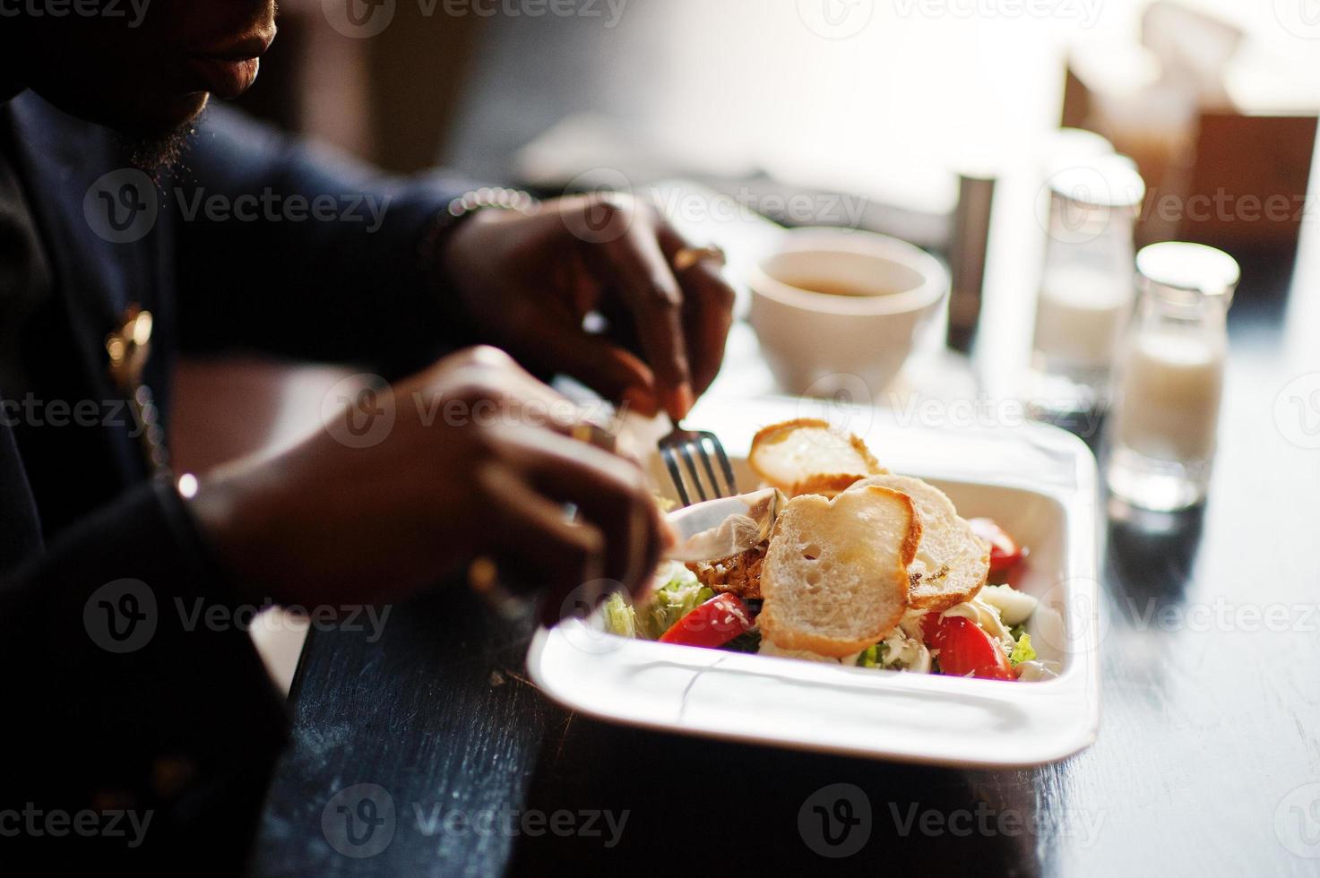 närbild händer på fashionabla afrikansk amerikansk man i kostym sitter på café och äter sallad. foto